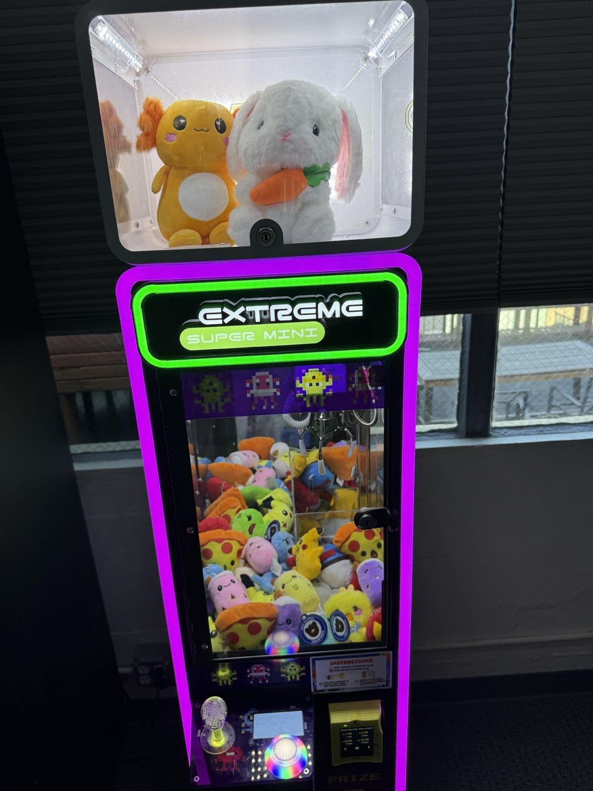 A purple-lit claw machine by a window with a city view, an orange plush and a white bunny in the display box on top