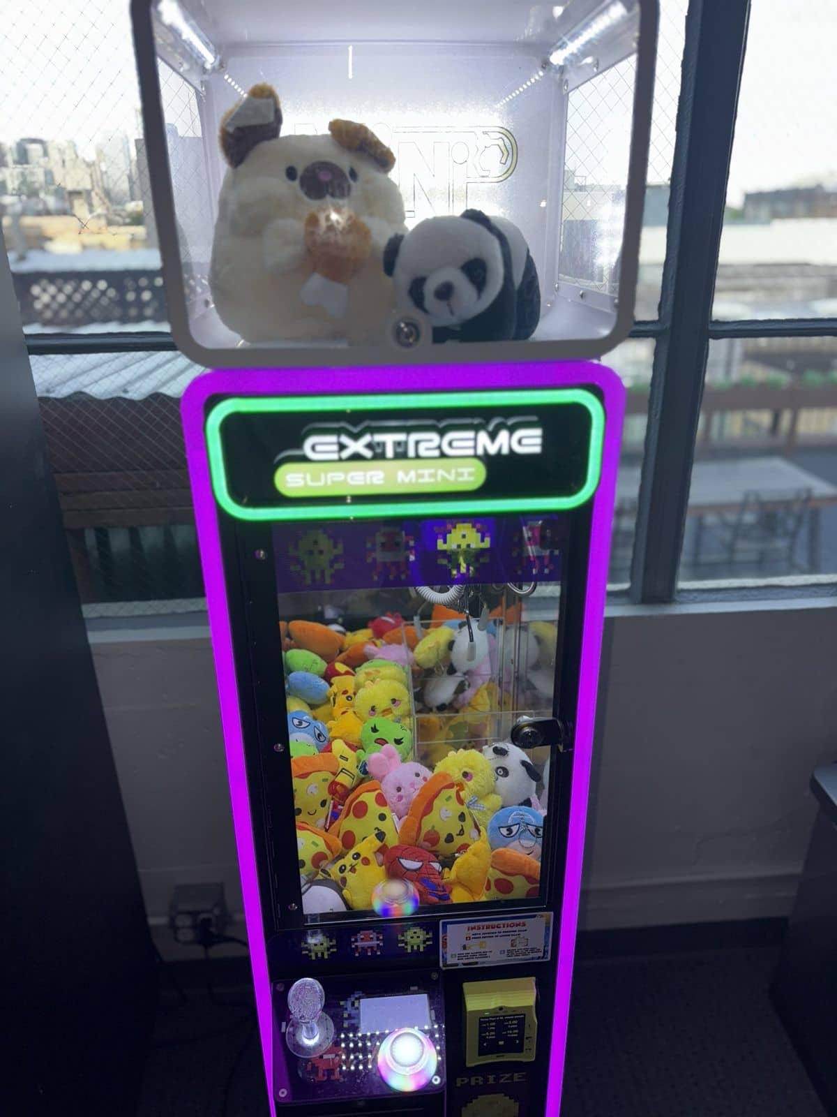 A purple-lit California Claw machine beside a tall window with a city view, a panda and a dog plush in the display box on top
