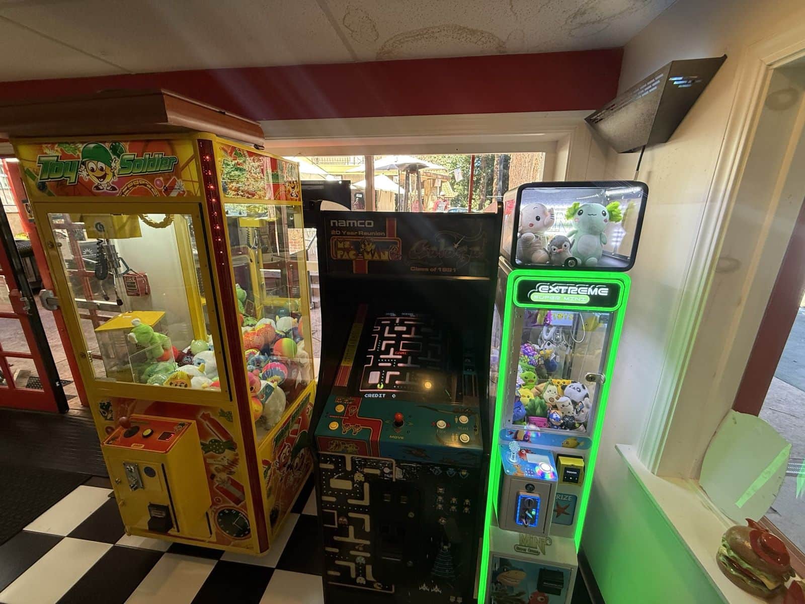 A green-lit claw machine next to a yellow crane machine and a classic arcade cabinet on a checkered floor