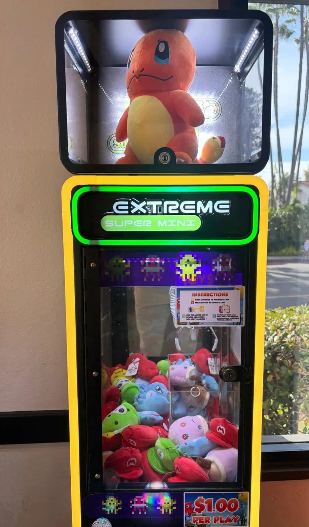 A claw machine beside a window with palm trees outside, holding Charmander, Poké Ball, and Mario cap plushies
