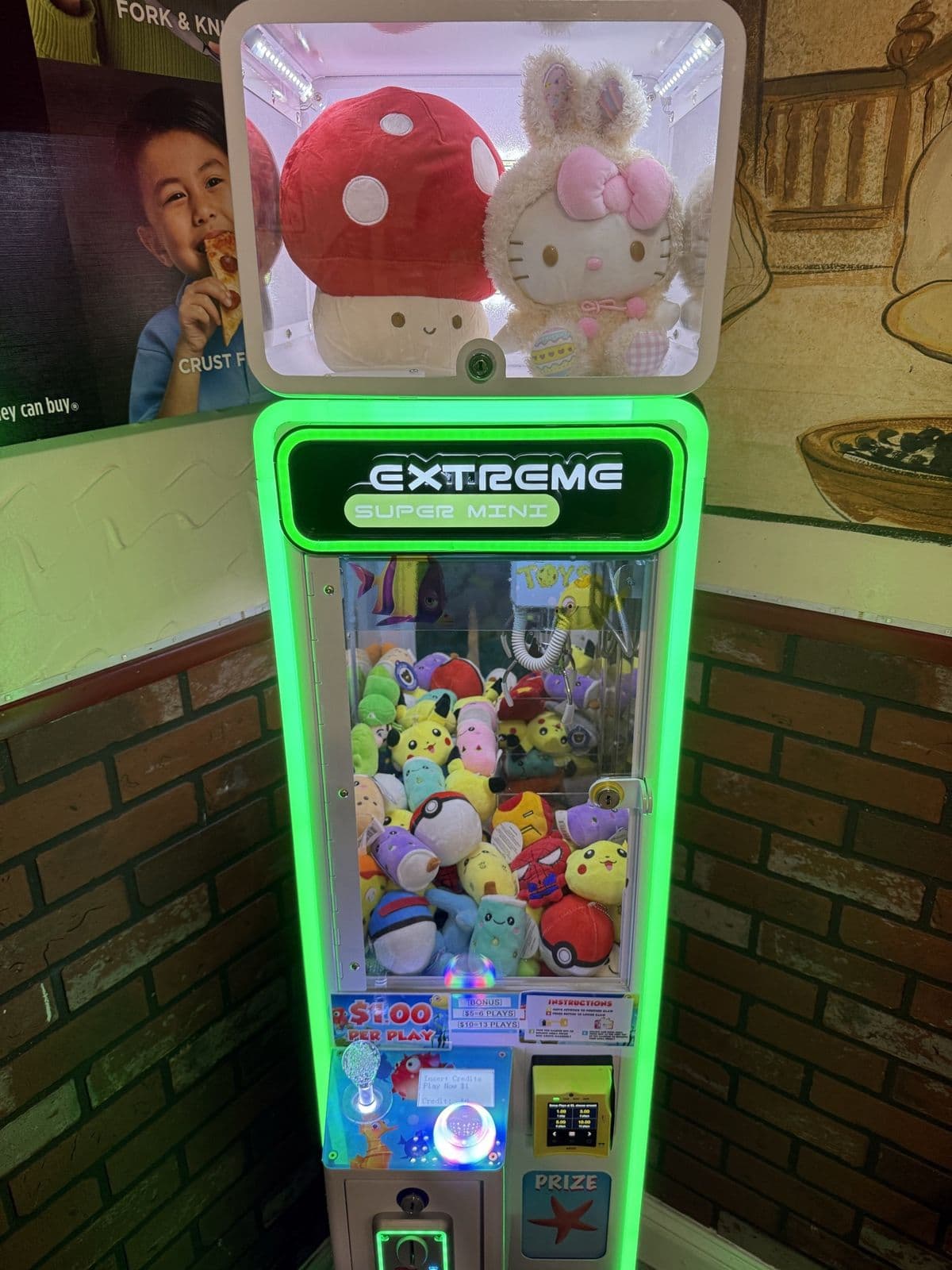 A green-lit California Claw machine in a pizzeria corner with plush in the display box on top
