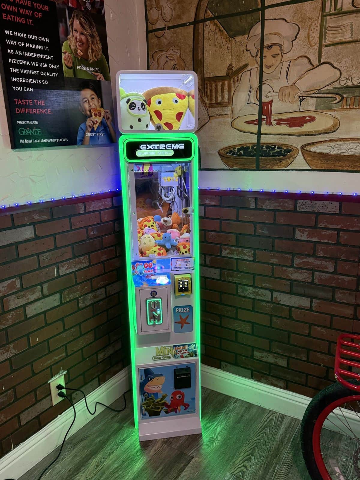 A green-lit claw machine in the corner of a pizzeria, brick wall and mural behind it