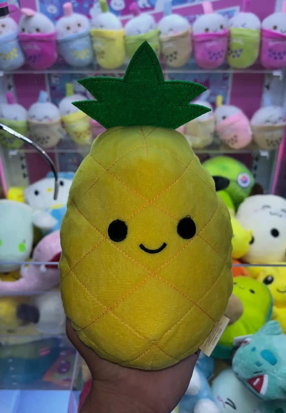 A hand holding a smiling pineapple plushie in front of a claw machine full of boba-cup plushies