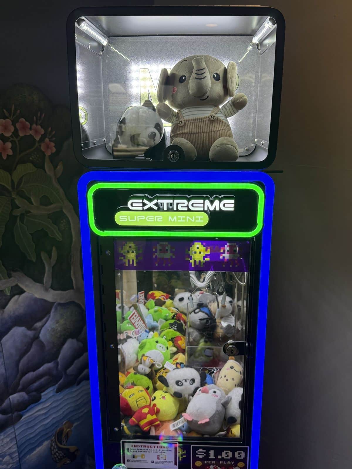 A blue-lit claw machine against a waterfall mural, an elephant plush in the display box on top