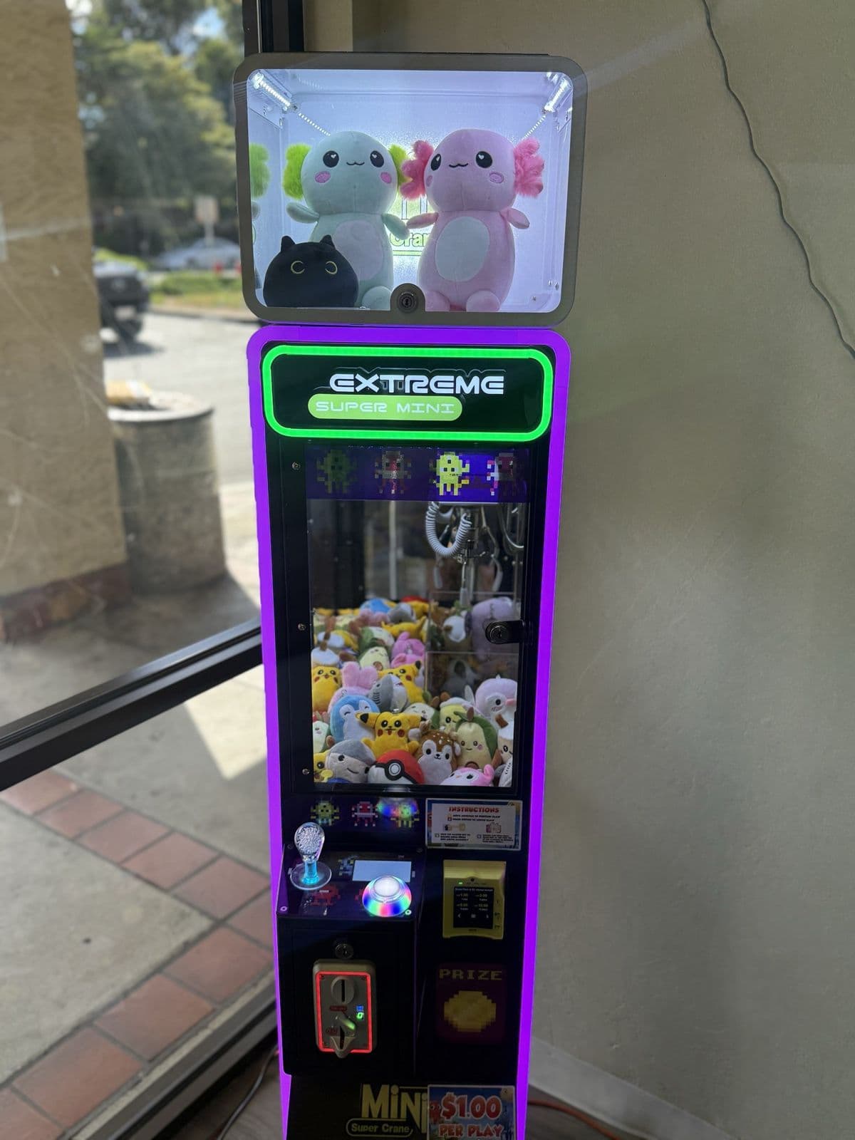 A purple-lit California Claw machine by a café window, two axolotl plushies on top