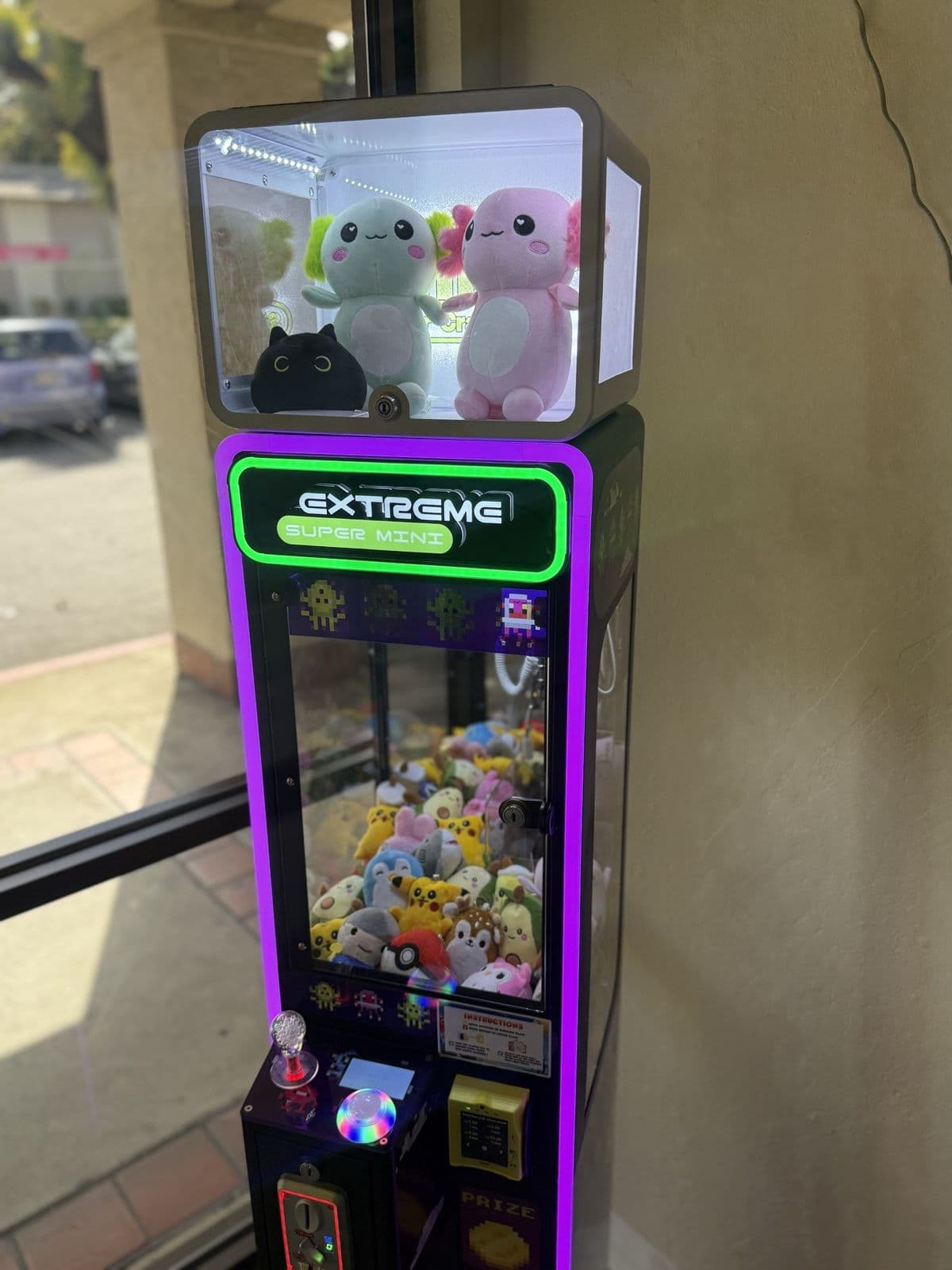 A purple-lit claw machine by a café window with two axolotl plushies on top